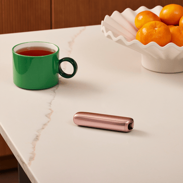 Ploom AURA Rose Gold diletakkan di meja kafe semasa rehat kopi, digayakan untuk penggunaan gaya hidup santai.