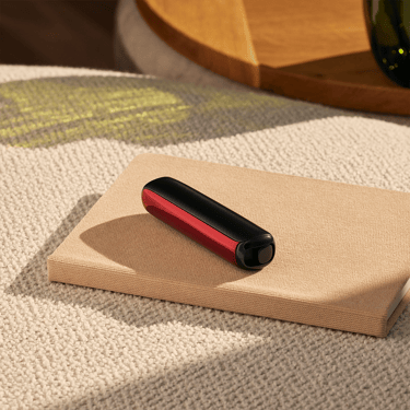 Ploom AURA dengan panel Lava Red diletakkan di atas meja, memperlihatkan reka bentuk modular dan kontras warna menarik dalam suasana gaya hidup.
