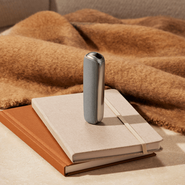 Ploom AURA with Lunar Silver Back Cover on a notebook stack, emphasizing smooth curves, minimalist metallic design, and refined modern aesthetics.