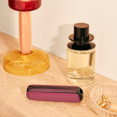 Ploom device with a Dark Orchid Back Cover placed on table beside a perfume, highlighting premium design and a polished, high-end lifestyle setting
