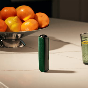 Peranti Ploom dengan Amazon Green Back Cover di atas meja berhampiran bakul buah, menggabungkan warna semula jadi dengan suasana gaya hidup hangat.
