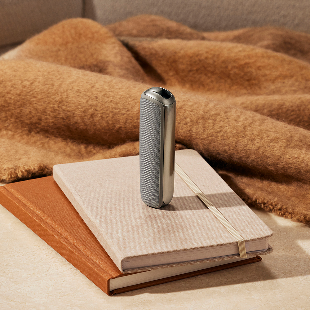 Ploom AURA with Lunar Silver Back Cover on a notebook stack, emphasizing smooth curves, minimalist metallic design, and refined modern aesthetics.

