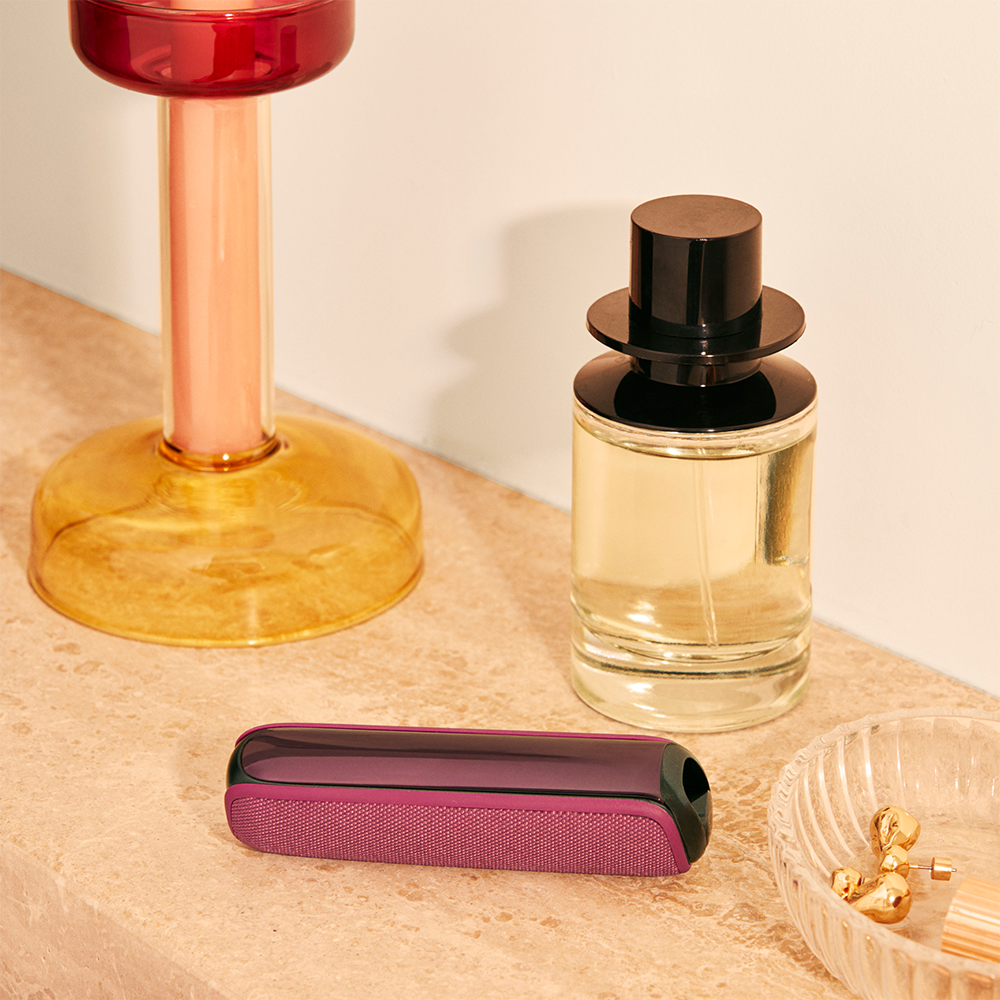 Ploom device with a Dark Orchid Back Cover placed on table beside a perfume, highlighting premium design and a polished, high-end lifestyle setting
