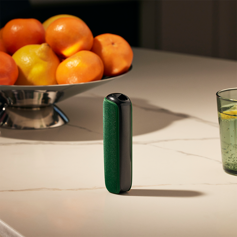 Ploom device with Amazon Green Back Cover on a table near a fruit basket, blending natural color with a warm, lived-in lifestyle scene
